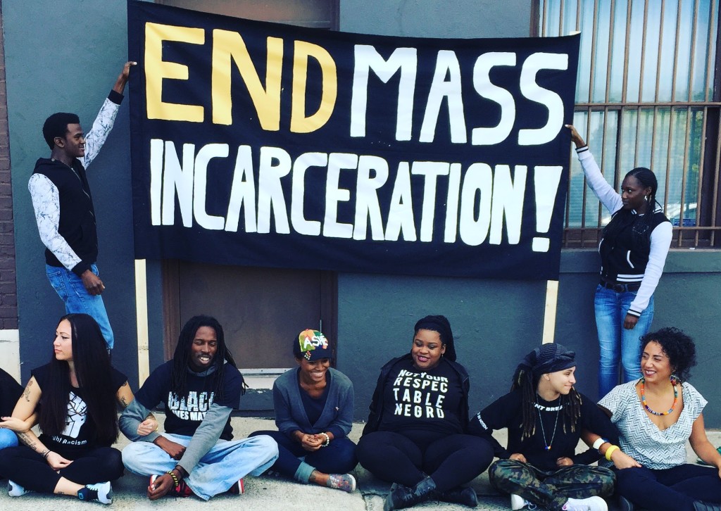 A multi-ethnic group of people pose around a large sign printed with "End Mass Incarceration!" in bold letters. They have their arms interlinked.